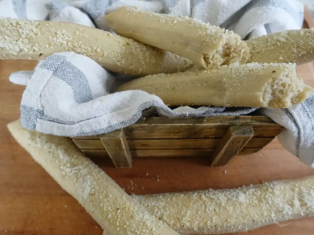 sourdough breadsticks in a tea towel lined basket with garlic parmesan topping with one broken in half so you can see how fluffy they are