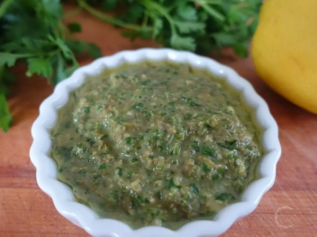 salsa gremolata in a small white dish on a wooden cutting board with the parsley and lemon used to make it behind it