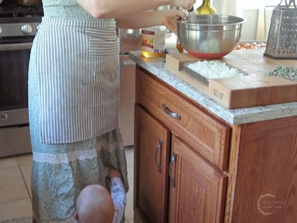 woman with an apron stirring food in a bowl in her kitchen with a baby tugging at her dress while shes cooking dinner