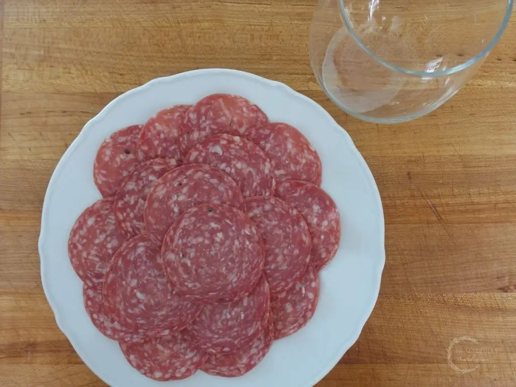 white plate with salami on it with a wine glass next to it showing what you need to make a salami rose