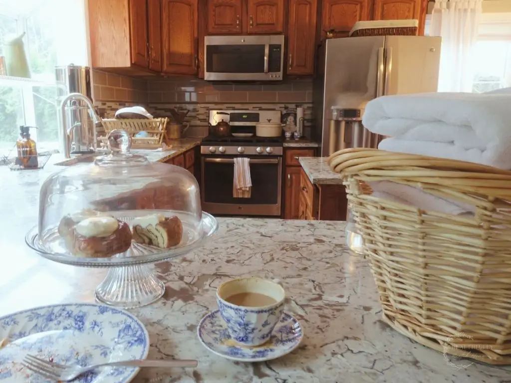 a laundry basket full of folded white towels sitting on top a granite countertop next to a cake stand with cinnamon rolls inside and blue and white china next to them. Showing how keeping pretty things in your home is one of the secrets to a clean house