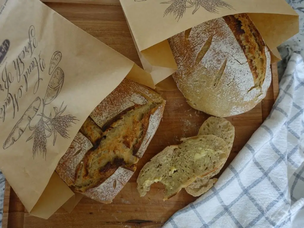 two loaves of sourdough bread in a brown paper bag one sliced with buttered slices next to it