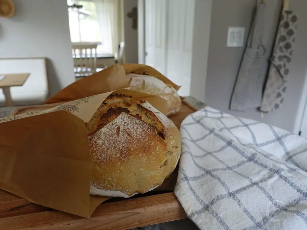 sourdough bread boule being stored in a brown paper bag placed on a wooden cutting board next to a white and blue striped tea towel
