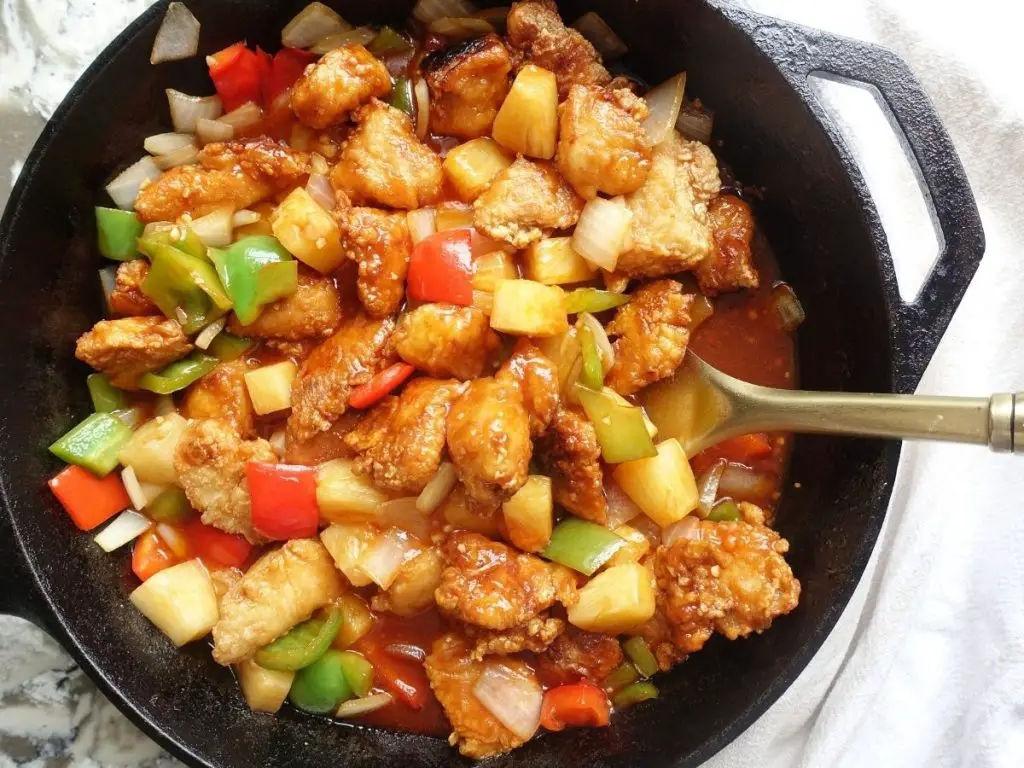sweet and sour chicken hong kong style in a cast iron skillet with a gold spatula resting inside