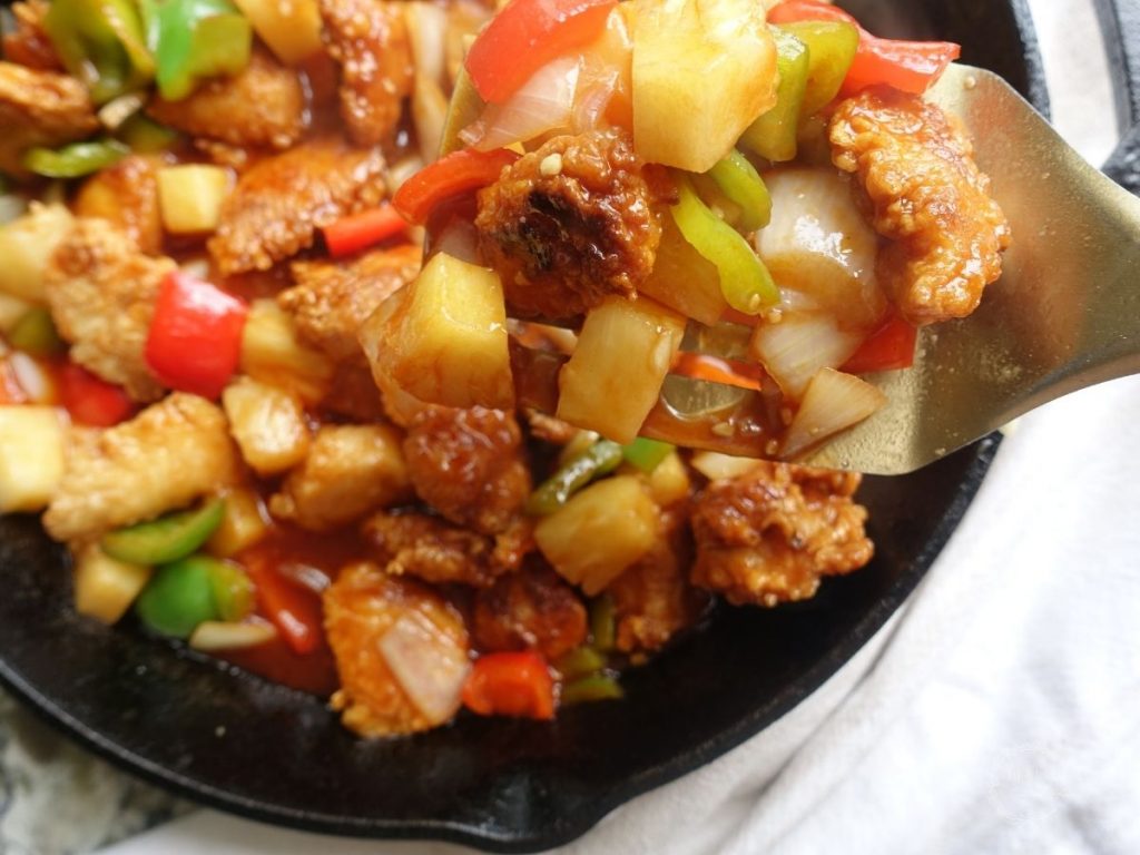 close up of sweet and sour chicken hong kong style in a cast iron skillet with a spatula holding up a scoop