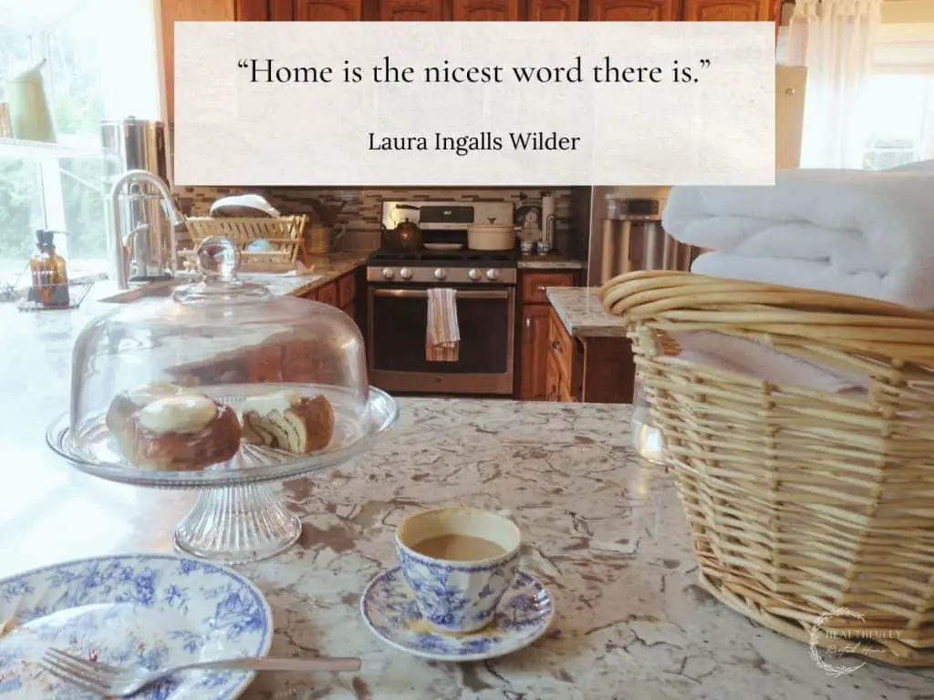 homemaking scene with a laundry basket full of white towels and a cake stand with cinnamon rolls inside and a cup of coffee on blue and white china displaying a homemaking quote