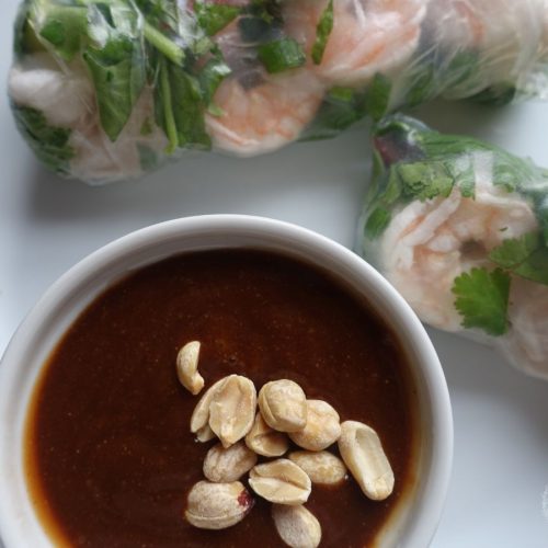 hoisin peanut sauce in a white bowl with chopped peanuts as the garnish and homemade spring rolls on the side