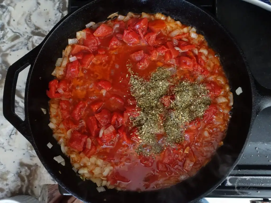 tomatoes and italian spices overtop onions simmering in a cast iron skillet