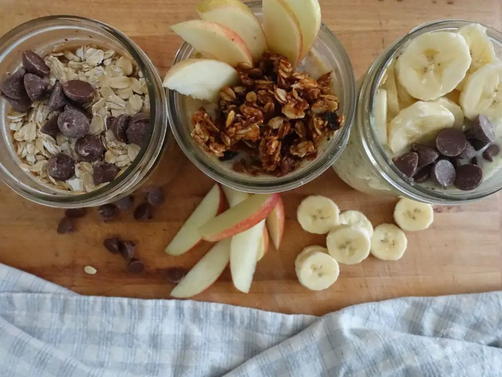 high protein overnight oats in 3 mason jars with 3 different flavors and toppings chocolate chips apples and bananas with a tea towel on the side for decor
