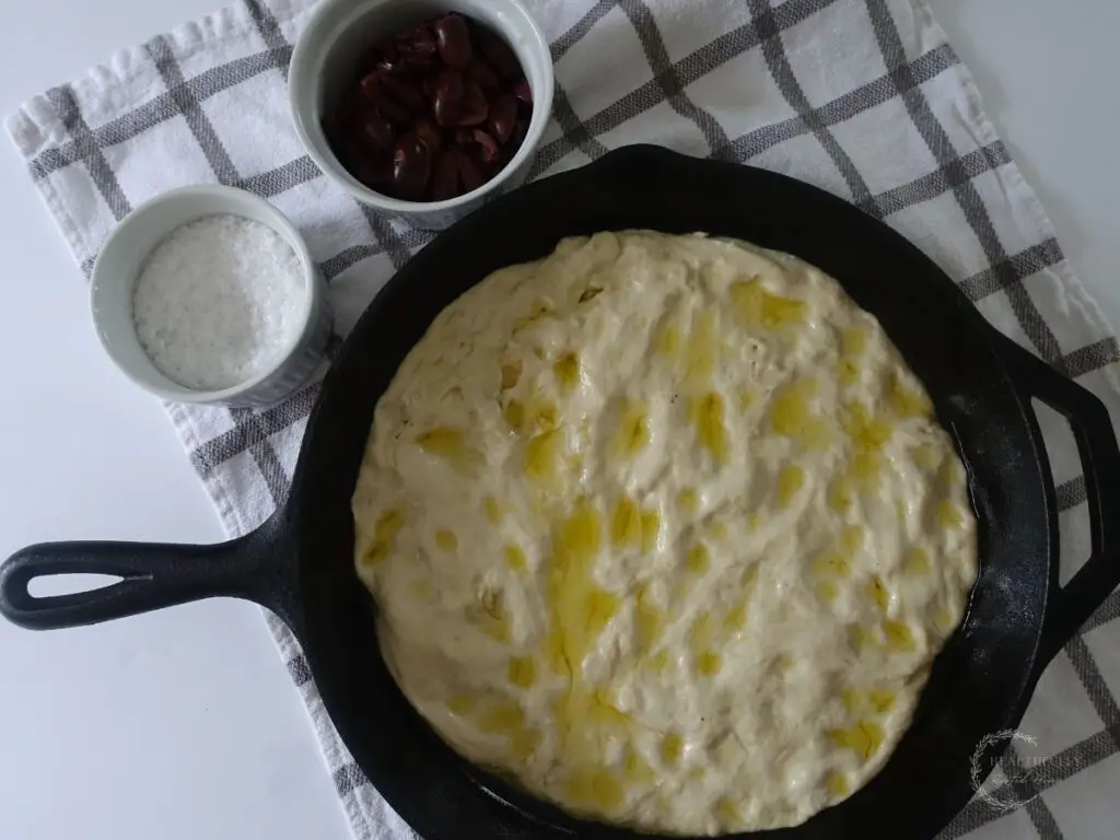 sourdough discard focaccia dough in a cast iron skillet after rising for a second time until puffy with the toppings on the side