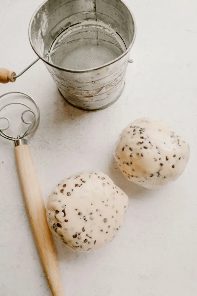 sugar cookie dough with chocolate chips rolled into balls and wrapped in plastic wrap with a danish dough whisk and a sifter next to it ready to be chilled